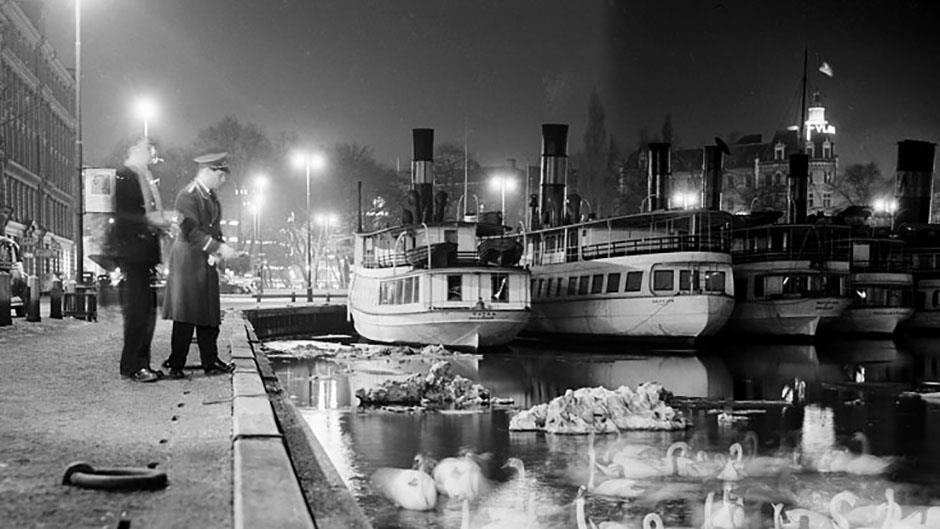 Två män matar svanar vid kajkanten. I bakgrunden Berzelii Park och till höger ångbåtar. Nybrokajen 28 januari 1955.