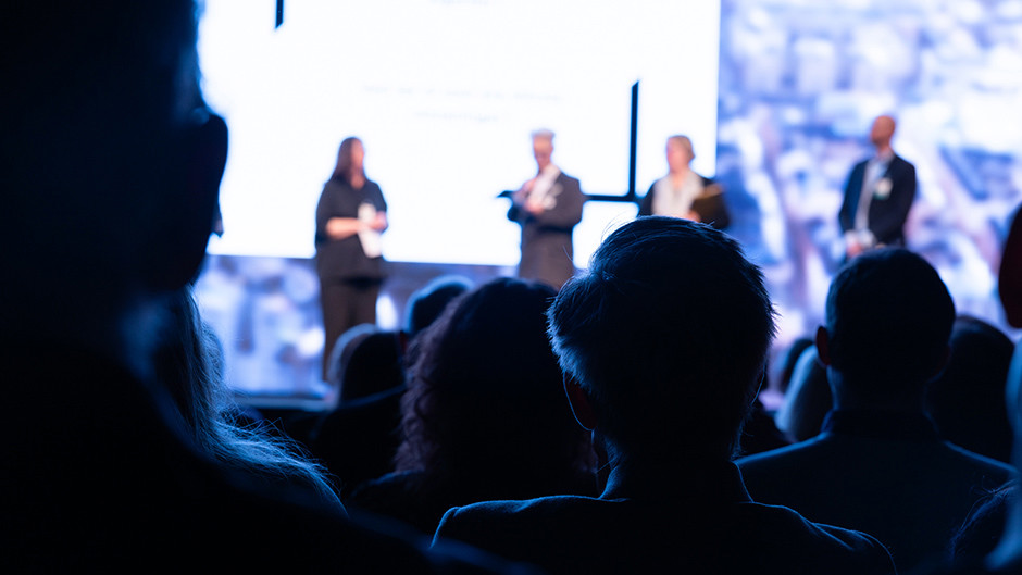 Bakhuvuden på åskådare i fokus. På scen står fyra personer ur fokus.
