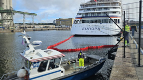 Två män i varselkläder lägger ut en läns i vattnet i Frihamnen. Kryssningsfartyg i bakgrunden.