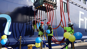Två personer i varselkläder håller i en jättestor elkontakt. Ett stort fartyg i bakgrunden.
