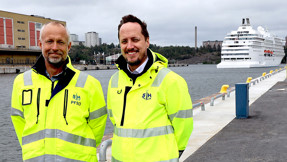 Två män i varseljackor på kajen i Frihamnen. I bakgrunden ett kryssningsfartyg.