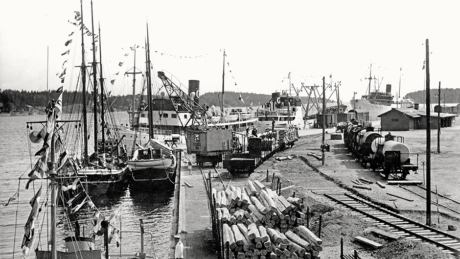 Nynäshamn år 1955. I förgrunden syns tågvagnar lastade med timmer. Längs kajen ligger Gotlandsbåtarna Gute, Gotland och närmast i bilden ligger trotjänaren Drotten från 1928.