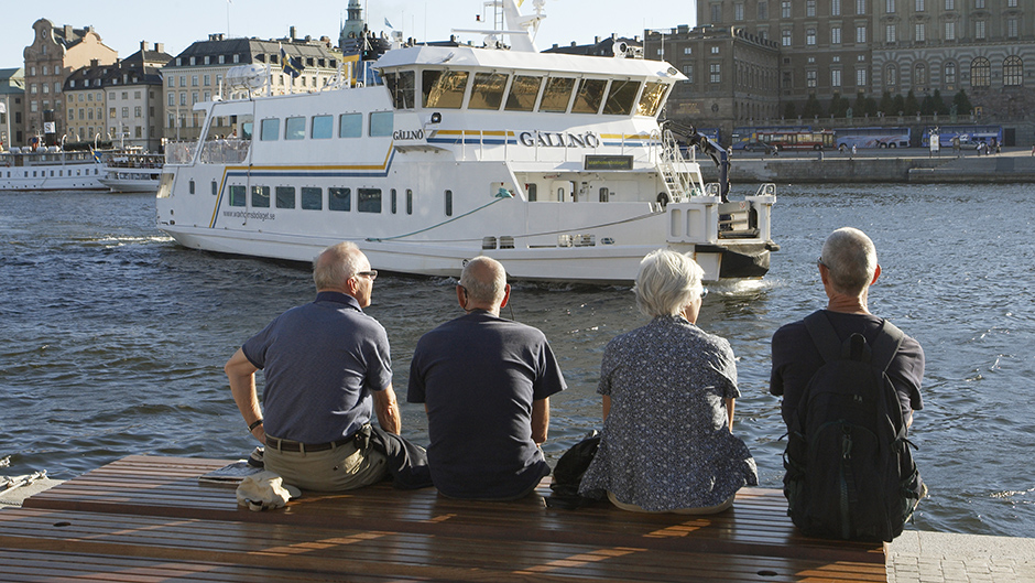 Fyra personer sitter på en bänk på Strömkajen med ryggen mot kameran och tittar på ett skärgårdsfartyg