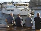 Fyra personer sitter på en bänk på Strömkajen med ryggen mot kameran och tittar på ett skärgårdsfartyg